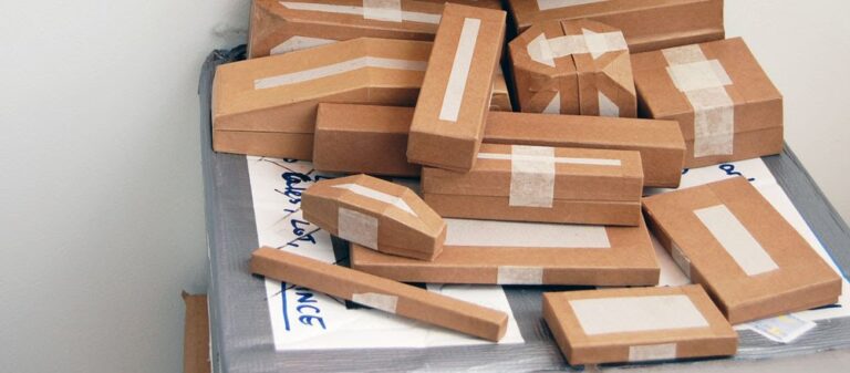A clutter of brown cardboard packages of various shapes and sizes, with white tape sealing them, haphazardly stacked on a surface partially covered with what appears to be white paper and gray duct tape along the edges, resembling an abstract artwork.