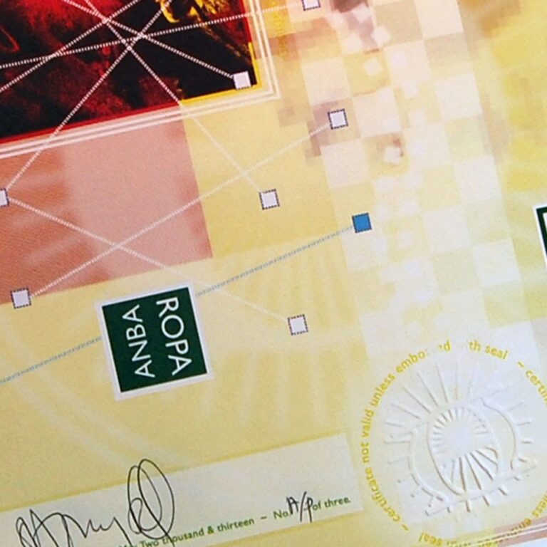 Close-up of a certificate displayed like an intricate art object, featuring a sun emblem, geometric patterns, and the text "ANBA ROPA." Signatures adorn the bottom and a subtle watermark pattern plays across the surface. The certificate appears to be printed on sturdy cardboard.