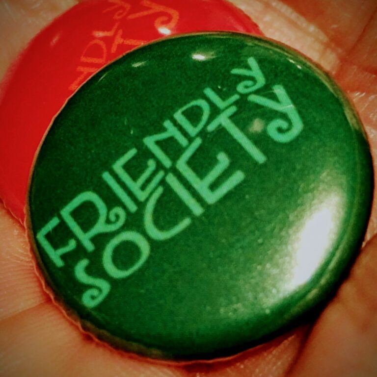 Close-up image of a green button pin with the words "FRIENDLY SOCIETY" written in a stylized font. Part of a red button pin with a similar design is partially visible in the background, both resembling artwork. The image suggests a theme of community or organization.