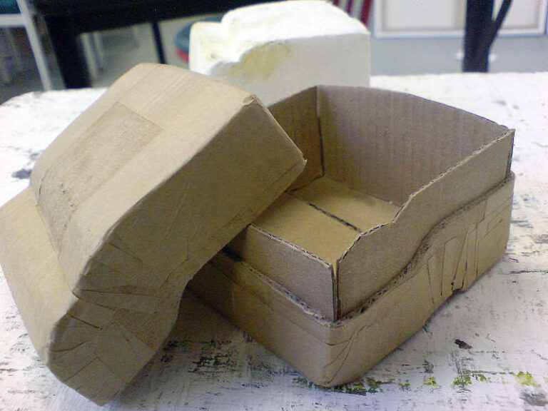 A handcrafted cardboard box, resembling an art object, sits on a worn white table. The roughly cut and assembled box features visible tape and folded edges. In the background, a blurred white object adds depth to the scene.