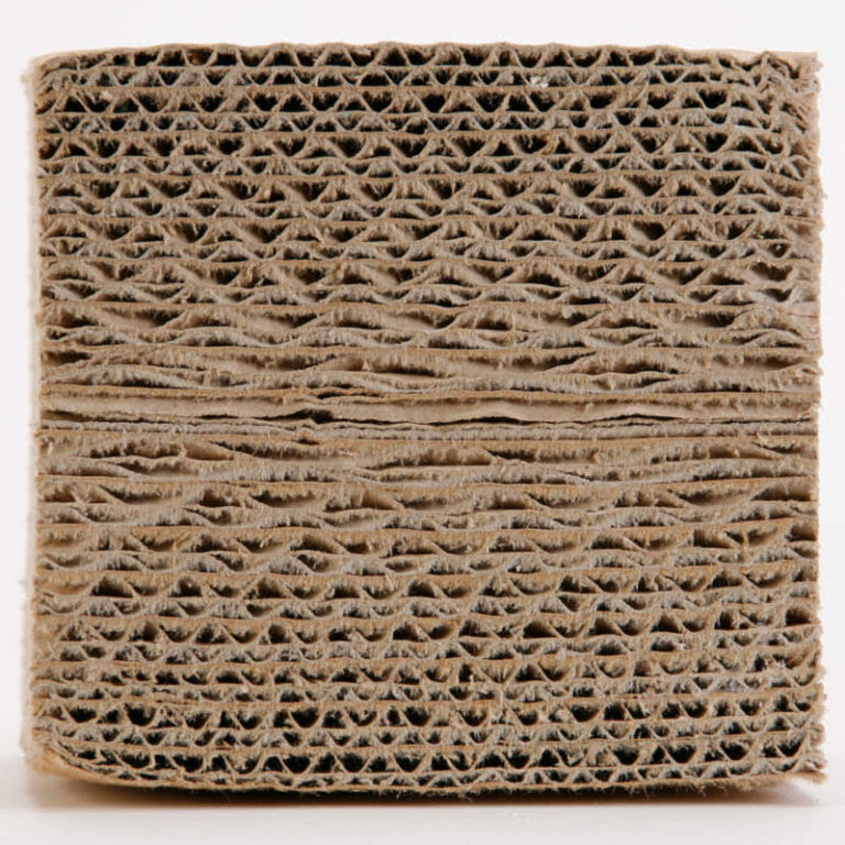 A close-up view of a thick, textured block of corrugated cardboard. The image highlights the intricate, uniform pattern of ridges and grooves typical of cardboard's layered, honeycomb-like structure. This worn and slightly compressed material transforms into an intriguing art object through its detailed simplicity.