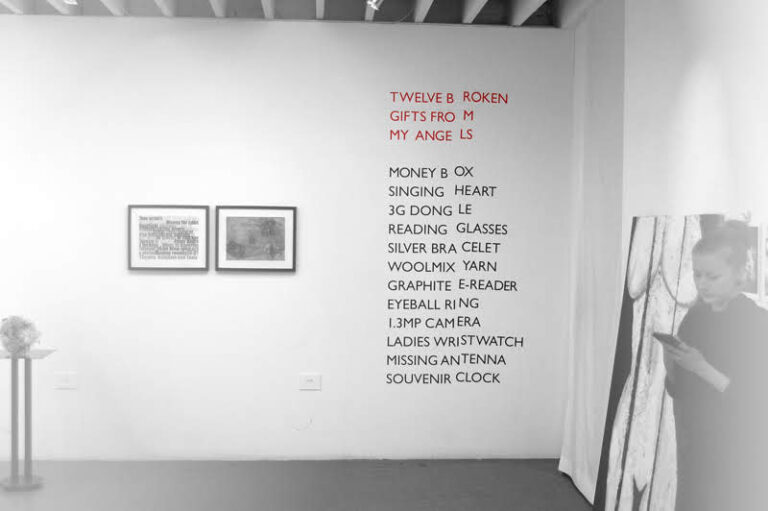 A minimalist room with white walls features a list of items in red text on the wall, titled "Twelve Broken Gifts From My Angels.” Near the list, a person stands using a smartphone. Two framed artworks are to the left, and a small side table with cardboard art objects beneath them completes the scene.