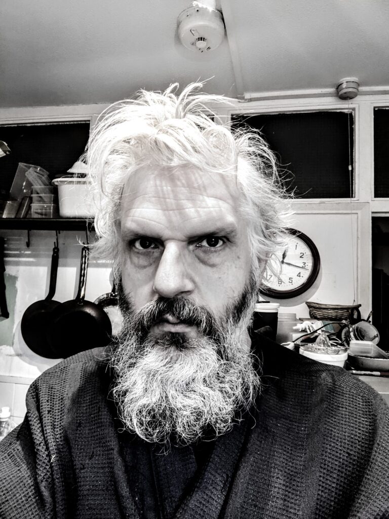 A black and white image of a man with tousled white hair and a thick, white beard, staring directly at the camera. He appears to be indoors with kitchen utensils and a clock visible in the blurred background. The lighting casts shadows on his face.