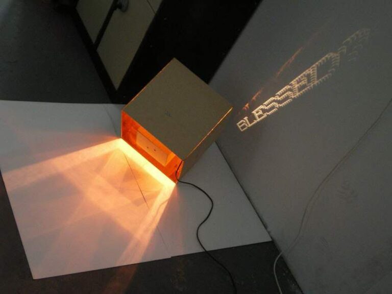 A cardboard box, transformed into an art object, is illuminated from the inside with a warm light, projecting a fragmented and abstract pattern onto a nearby wall. The box is placed on a white surface and has an electrical cord plugged into it.