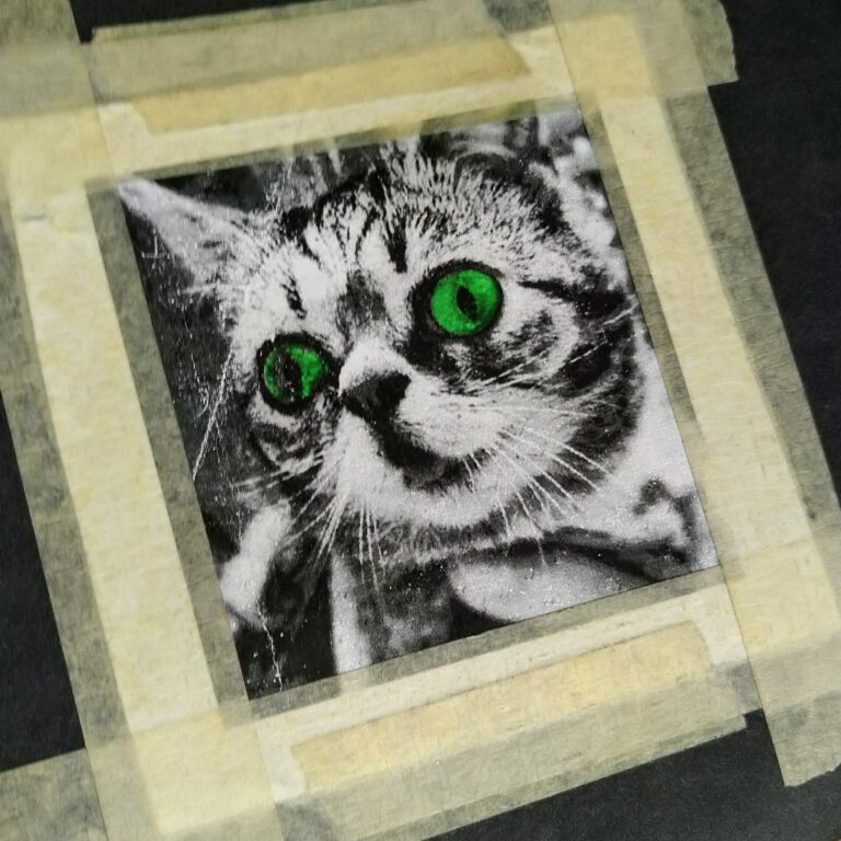 A close-up, black-and-white photograph of a cat with vivid green eyes framed by masking tape on a dark background. The cat's expression appears curious and slightly surprised. The masking tape forms a border around the photo.