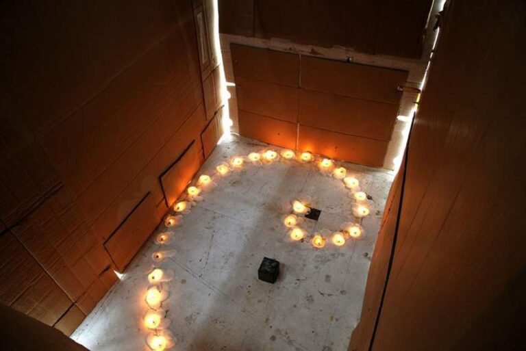 A room made of cardboard walls and ceiling, illuminated by small candles arranged in the shape of a question mark on the floor, serves as a unique artwork. Light filters through gaps in the cardboard, adding a warm glow to this intriguing art object's space.