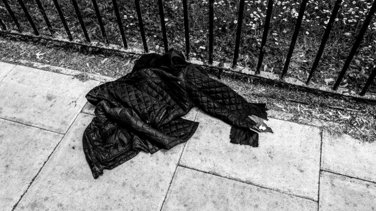 A black quilted jacket lies abandoned on a pavement next to a metal fence. The ground is a mix of concrete slabs and pebbles, with grass and small flowers on the other side of the fence. The image is in black and white.