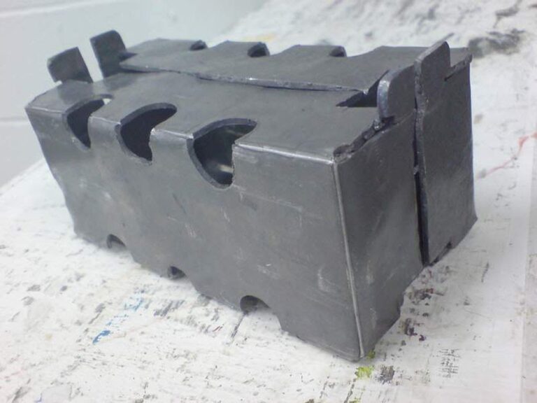 A block of dark gray modeling clay shaped to resemble a hollow rectangular prism with wavy edges and several circular cutouts on its sides. The surface shows a slightly textured appearance, indicative of manual shaping. This unique artwork rests on a white, slightly messy table.