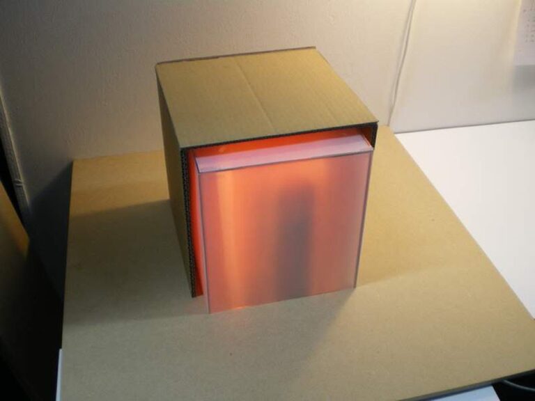 A small cardboard box with an open front sits on a tabletop, resembling a captivating art object. Inside the box, a semi-translucent pink material is illuminated, possibly a light source or display. The surrounding area is well-lit, showcasing this unique artwork as the focal point.