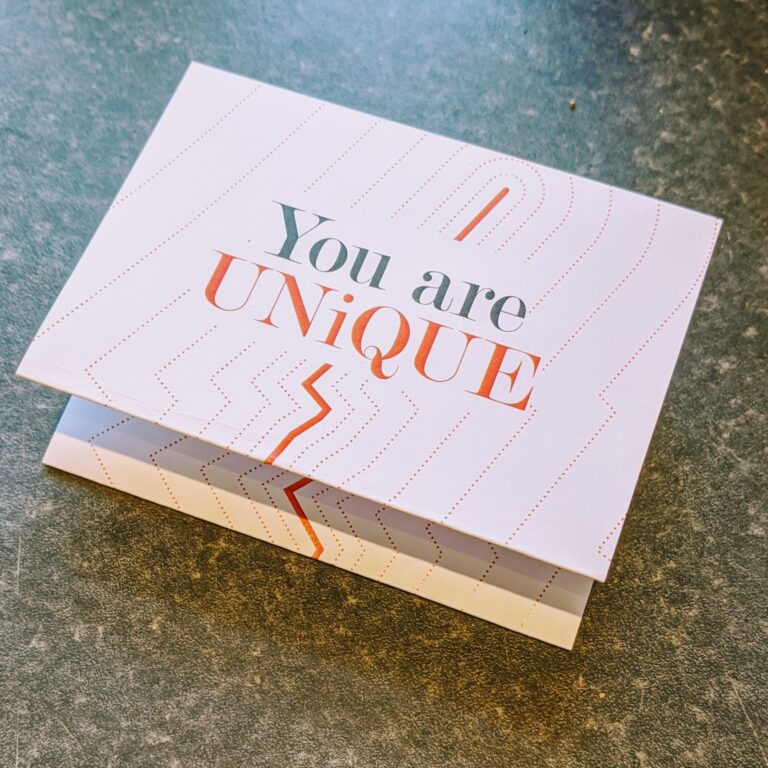 A folded greeting card on a grey surface with the message "You are UNIQUE" printed in bold letters. The card has a minimalist design with subtle line patterns and an orange and grey color scheme.